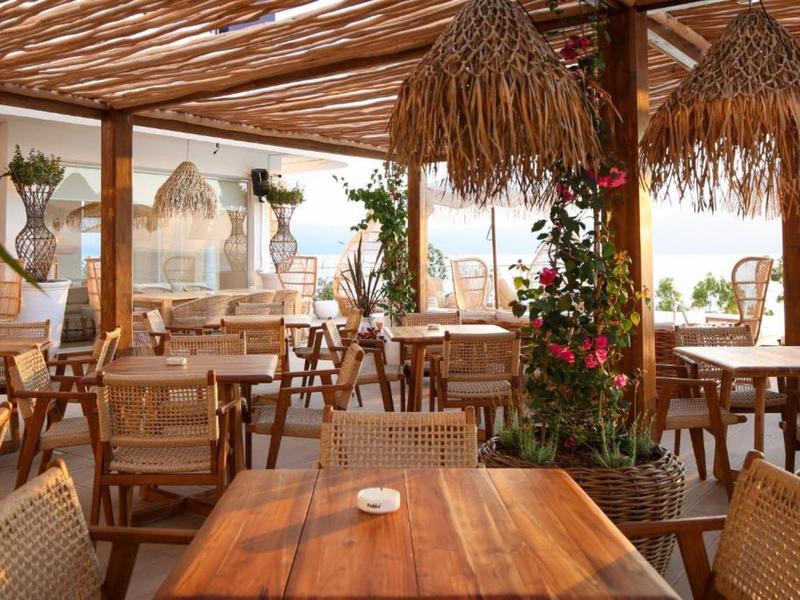 Hell eingerichtetes Restaurant mit Holztischen, Korbsesseln und Pflanzen unter einem geflochtenen Dach.