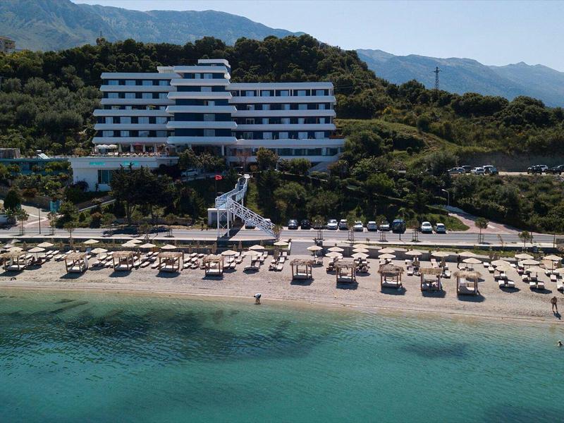 Großes Hotel am Hang mit Strand, Sonnenliegen und klarem Meer im Vordergrund.