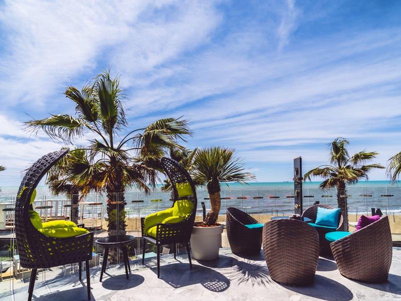 Strandterrasse mit Sitzgelegenheiten und Palmen vor blauem Himmel und ruhigem Meer