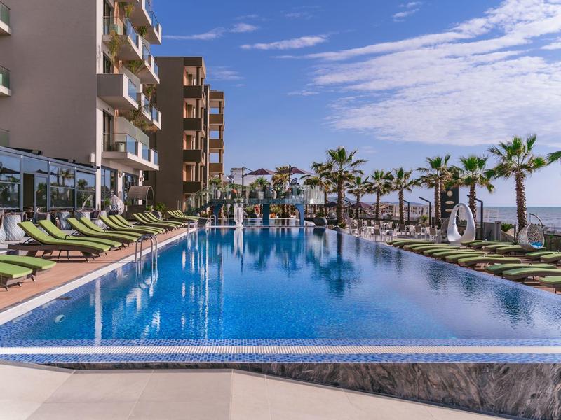 Moderner Outdoor-Pool mit Sonnenliegen und Palmen in einem Hotelresort bei sonnigem Wetter.