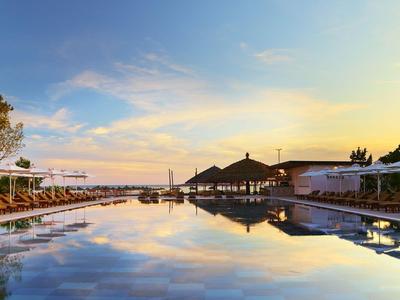 Vue sur une piscine calme avec des parasols et des arbres au coucher du soleil.