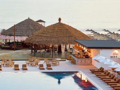 Strandresort mit Pool, Liegestühlen, Sonnenschirmen und großen Strohdächern am Meer.