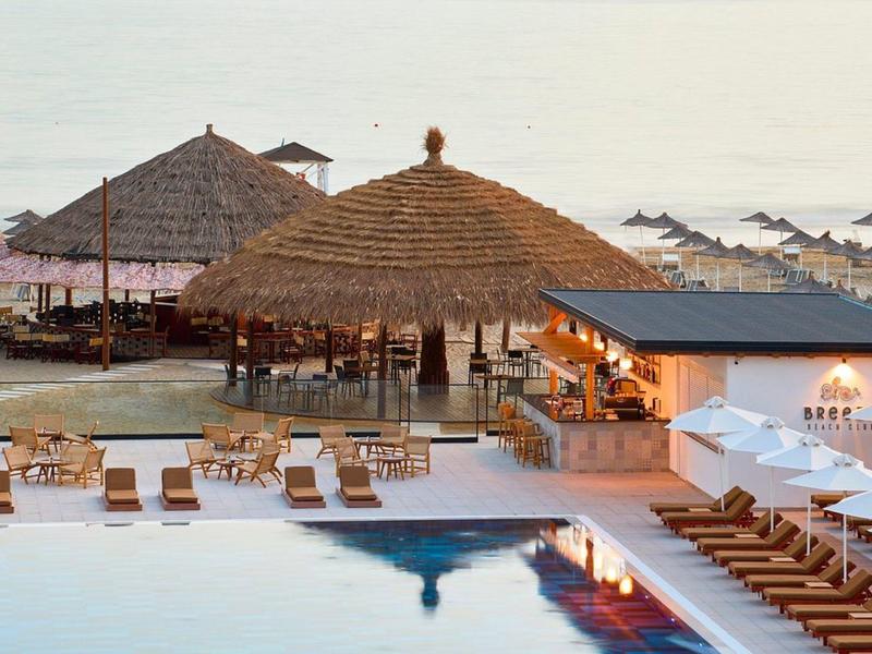 Strandresort mit Pool, Liegestühlen, Sonnenschirmen und großen Strohdächern am Meer.