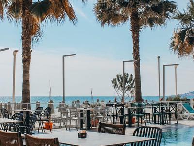 Espace extérieur moderne avec chaises et tables près d'une piscine, palmiers et vue sur la mer en arrière-plan.