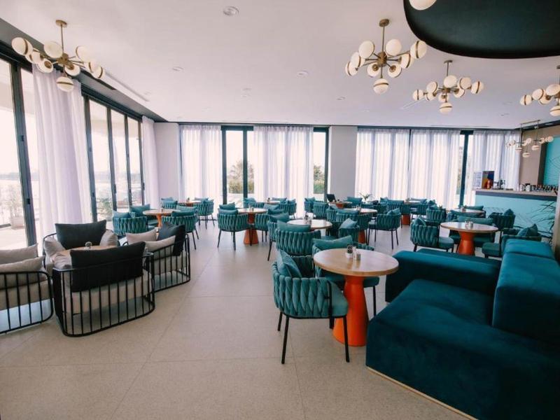 Heller, moderner Gastraum mit blaugrünen Sesseln, runden Tischen und großen Fenstern, viel Tageslicht.