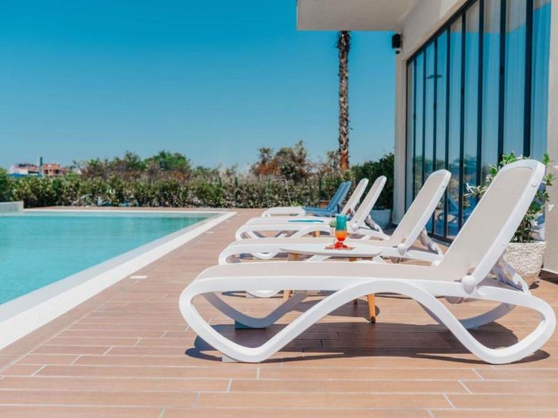 Reihe weißer Liegestühle neben klarem Pool mit glasener Hauswand und blauem Himmel.