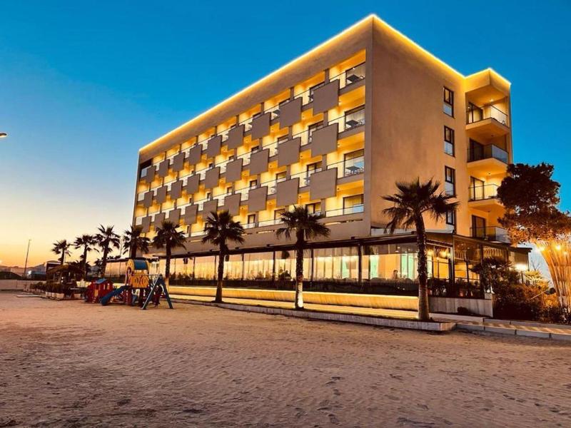 Modernes Hotelgebäude mit beleuchteten Balkonen, Palmen und Sandstrand bei Sonnenuntergang.