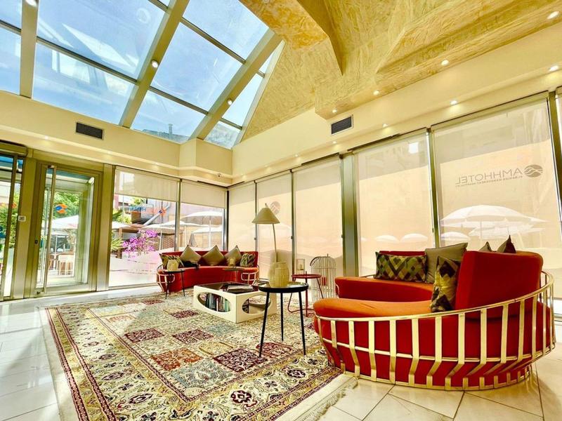 Heller Raum mit großen Fenstern, roten Sesseln, Teppich und gläsernem Dach.