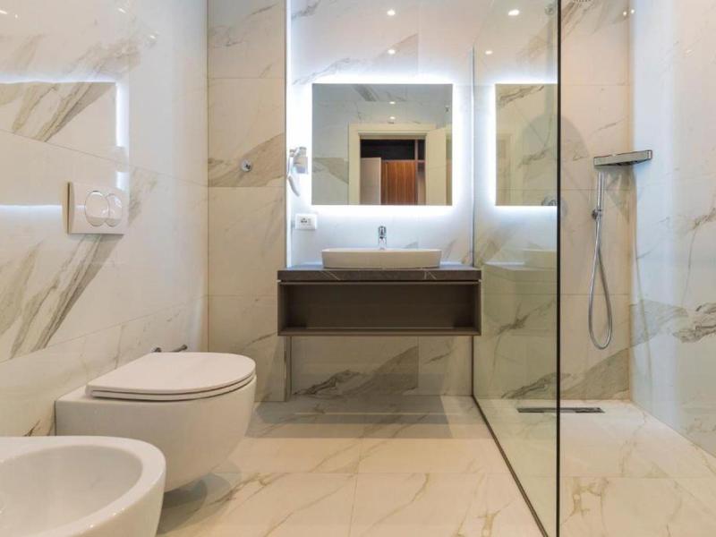 Modernes Badezimmer mit Marmorfliesen, beleuchtetem Spiegel, Toilette, Bidet und Glasduschkabine.