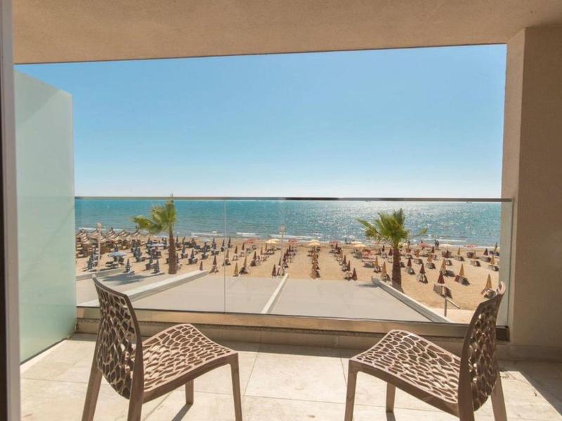 Blick von Balkon mit zwei Stühlen auf Strand mit Sonnenschirmen, Palmen und blauem Meer am Horizont.