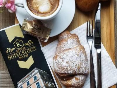 Kaffeetasse mit Cappuccino, zwei Brötchen, Croissant mit Puderzucker, Besteck und Hotel-Flyer auf Holzbrett.