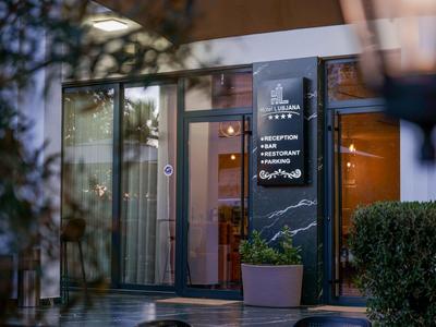 Entrée d'un hôtel avec panneau illuminé et plantes, entourée de verre et de végétation verte.