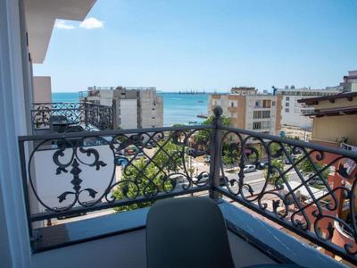 Balcon avec rampe en métal orné, vue sur des immeubles et la mer en arrière-plan.