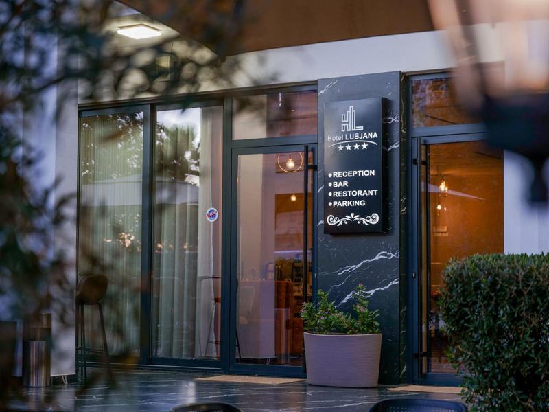 Vstup do hotelu se skleněnou fasádou, cedulí a květinami v květináčích.