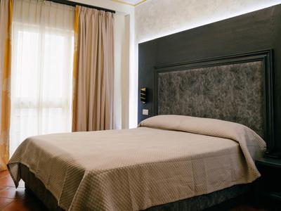 Hotel room with double bed, beige bedspread, dark headboard, and light curtains at window.
