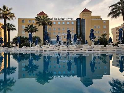 Ein Hotel mit gelber Fassade, Palme und blauem Pool, Spiegelung im ruhigen Wasser.