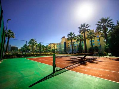 Campo da tennis con superficie rossa e linee verdi, circondato da palme e hotel al sole.