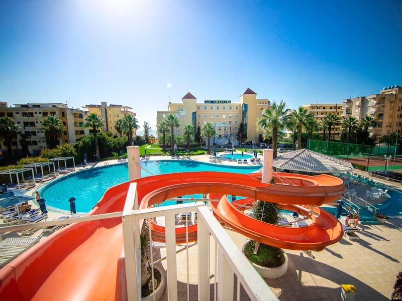 Duży basen hotelowy z czerwoną zjeżdżalnią otoczony leżakami i budynkami pod czystym niebem.
