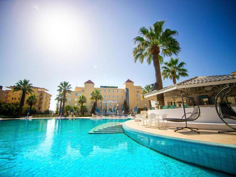 Duży basen z leżakami i palmami przed budynkiem hotelu pod słonecznym niebem.