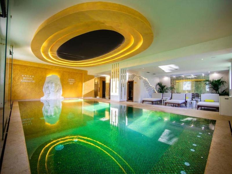 Basen kryty z zieloną wodą, zakrzywionym oświetleniem sufitowym i nowoczesnymi meblami w hotelu.