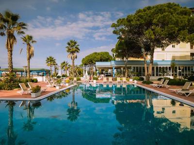 Großer Pool mit Palmen und Liegestühlen vor mehrstöckigem Hotel unter blauem Himmel am Meer.