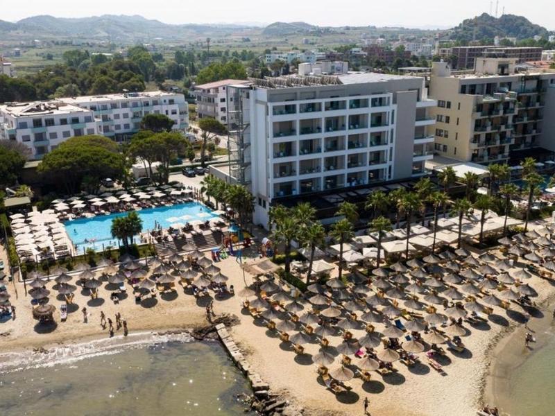 Complesso alberghiero sulla spiaggia con piscine, lettini e ombrelloni, circondato da montagne.