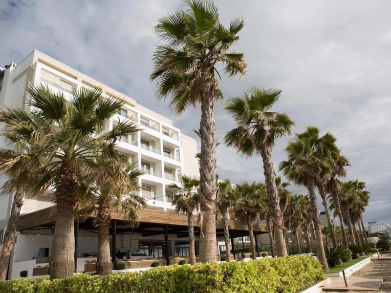 Edificio dell'hotel con balconi bianchi e fila di palme sotto un cielo nuvoloso.
