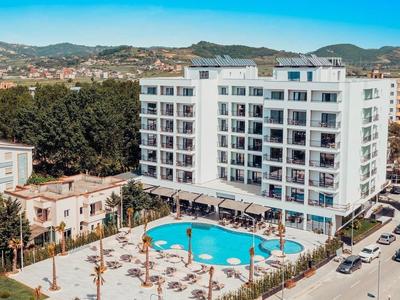 Complesso alberghiero moderno con piscina, circondato dalla città e dalle montagne sotto un cielo limpido.