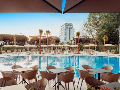 Pool mit blauer Wasserfläche, beige Stühle und Tische unter Sonnenschirmen, Hochhaus im Hintergrund.