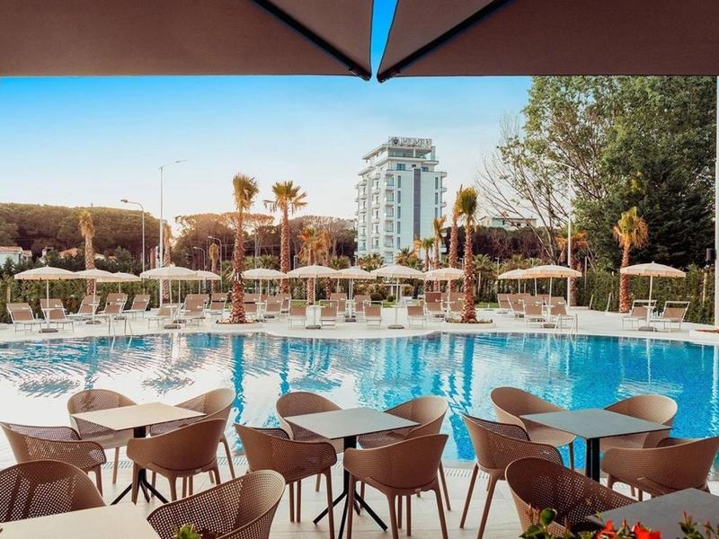 Vista di una piscina con lettini e tavoli all'aperto sotto un cielo limpido.