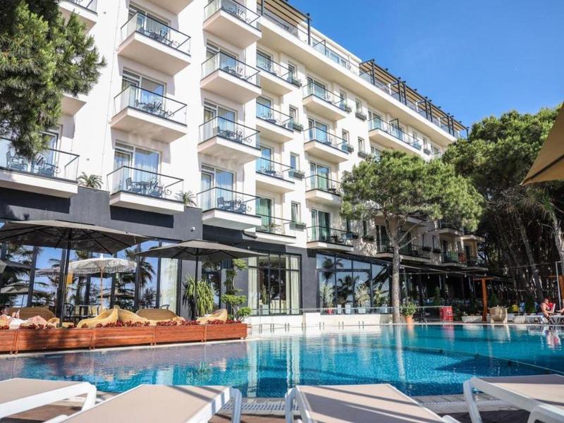 Hotel di cinque piani con piscina all'aperto e lettini sotto un cielo azzurro.