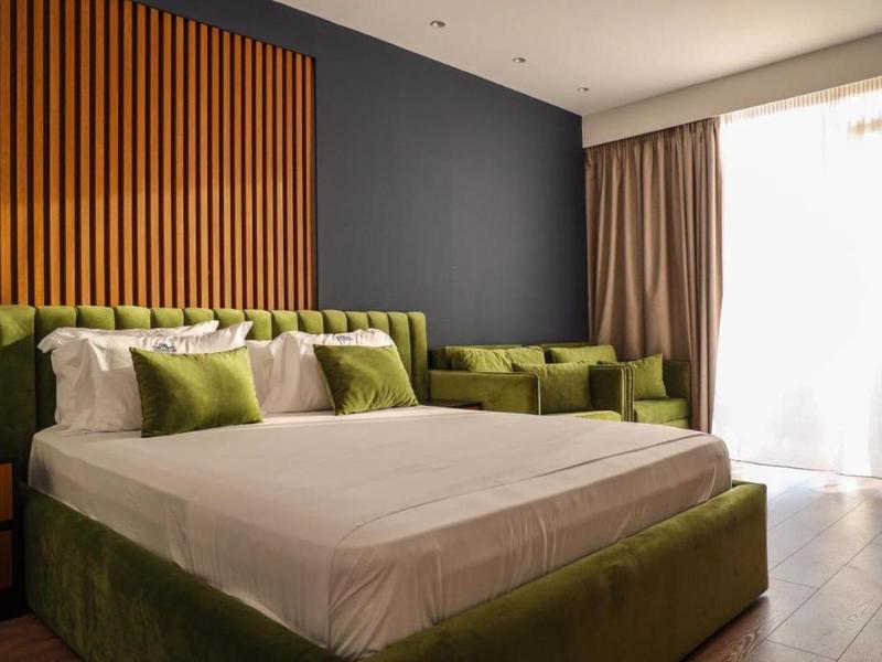 Camera d'albergo moderna con letto grande, area salotto verde e luce naturale da finestre a tutta altezza.