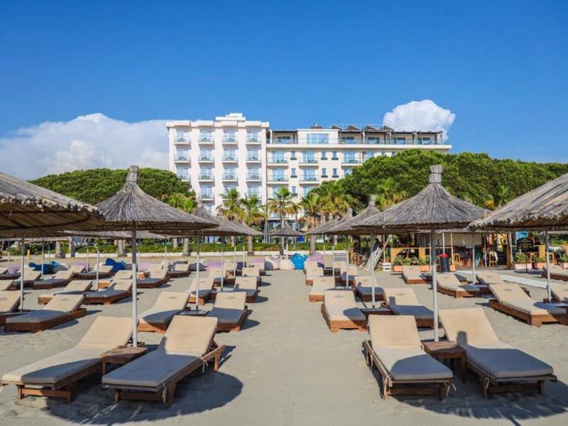 Spiaggia con lettini e ombrelloni davanti a un hotel di più piani sotto un cielo limpido.