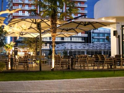 Außenbereich eines Cafés mit Tischen, Stühlen und Sonnenschirmen bei Abendlicht und Hochhaus im Hintergrund.