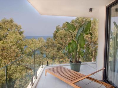 Balcone con lettino in legno, piante e vista su alberi e mare sotto un cielo limpido.