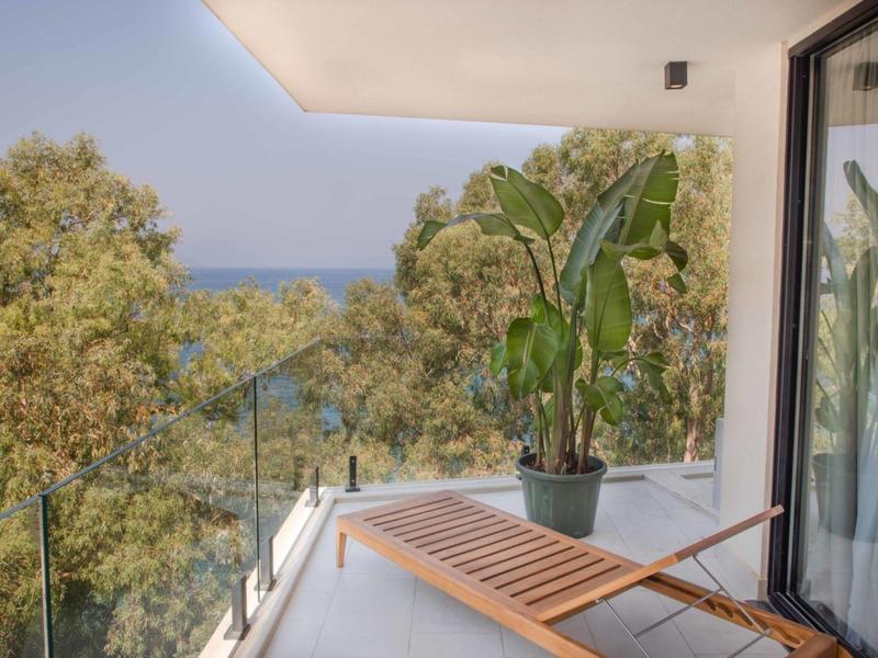 Balcone con lettino in legno, piante e vista su alberi e mare sotto un cielo limpido.