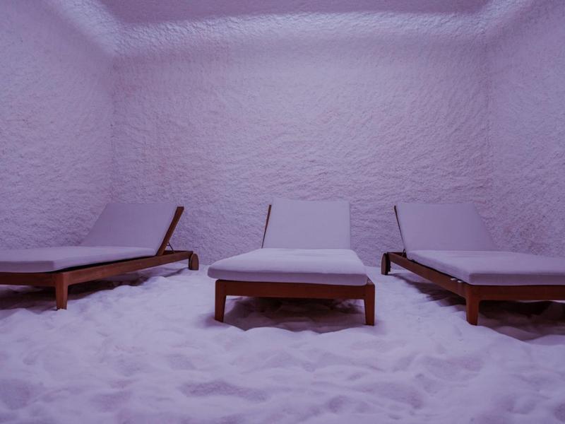 Tre lettini in legno con cuscini in una stanza con pareti e pavimento bianchi simili a sale.