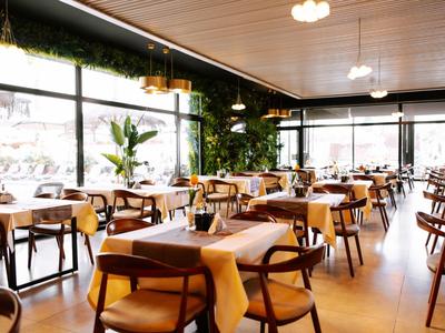 Restaurant lumineux avec tables en bois, chaises, grandes fenêtres et de nombreuses plantes décoratives.