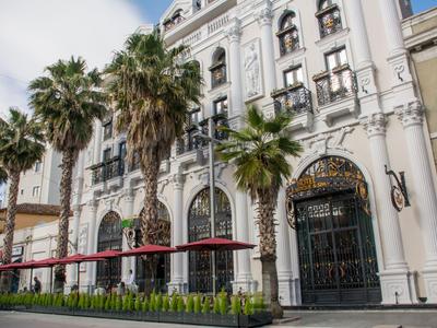 Historisch hotelgebouw met palmbomen en rode parasols bij de ingang.