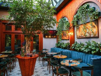 Interieur van een restaurant met bloeiende boom, blauw bankje en hangende planten aan oranje muur