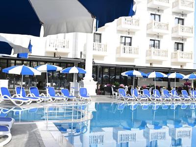 Piscina azul con tumbonas blancas y sombrillas azules frente a un edificio de hotel blanco.