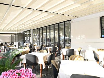 Restaurante cubierto y luminoso con mesas y sillas blancas junto a grandes ventanas de vidrio.