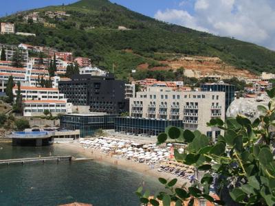 Strandbereich mit Sonnenschirmen und Liegen vor Gebäuden am Hang, umgeben von grünem Berg.