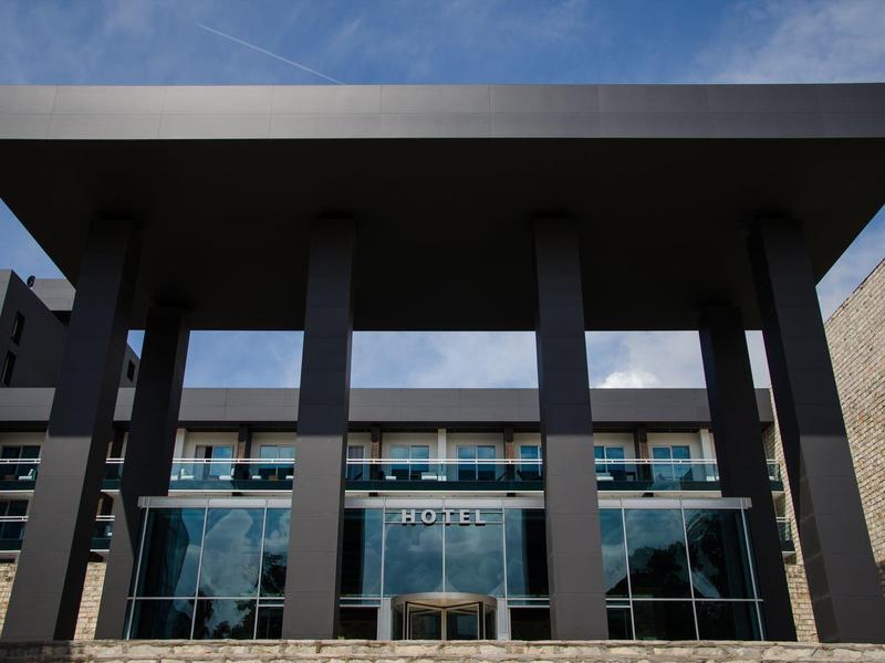 Edificio moderno con grandes ventanas y cuatro columnas oscuras en la entrada bajo cielo azul.