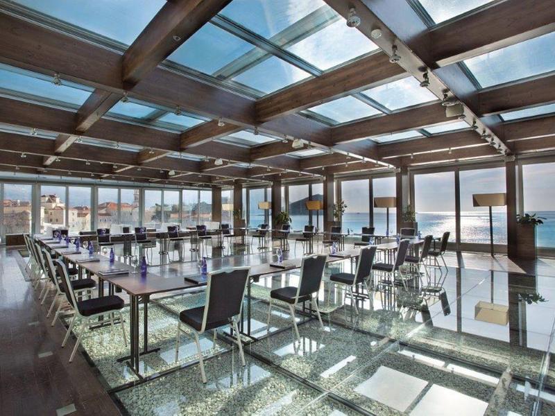 Restaurante luminoso con techo de vidrio y vista al mar, mesas con sillas sobre suelo de vidrio.