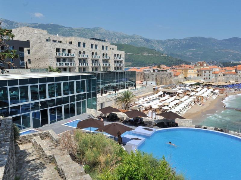 Hotel de lujo con piscina infinita y tumbonas junto al mar frente a montañas.