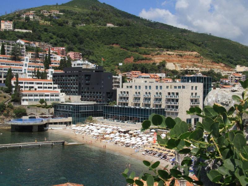 Complejo hotelero en la costa con piscina, sombrillas y colina al fondo.