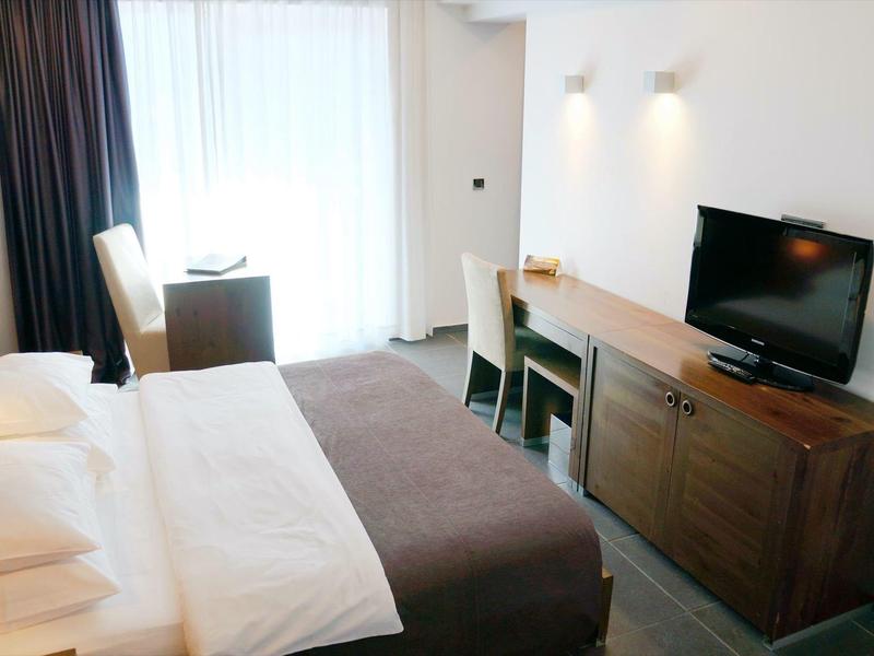 Habitación de hotel luminosa con cama, escritorio, televisor y ventana grande con cortinas.