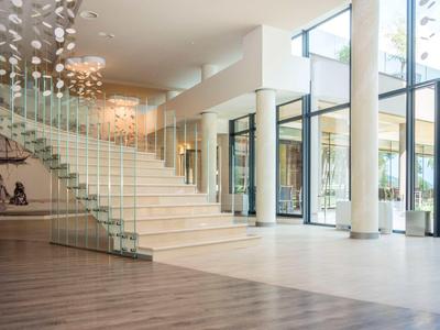 Área luminosa del vestíbulo del hotel con grandes ventanales, escalera con barandilla de vidrio y iluminación moderna.