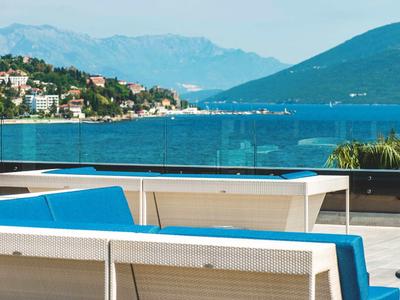 Blick von einer Terrasse mit Sitzgelegenheiten auf ein Meer und Berge im Hintergrund.